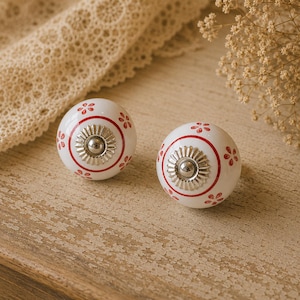 May include: Two white ceramic cabinet knobs with red floral accents and a silver-colored center. The knobs are round and have a decorative design, perfect for adding a touch of elegance to furniture or cabinetry.