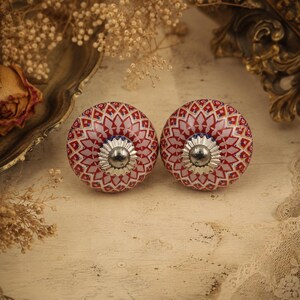 May include: Two decorative cabinet knobs. Each knob is round with a red and white geometric pattern. The knobs have a silver-coloured base and a blue accent. The knobs are on a light-coloured surface.