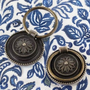 May include: Two antique-style metal drawer pulls with a circular design and a ring handle. The pulls have a dark bronze finish and intricate floral details.