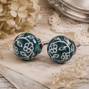 May include: Two teal cabinet knobs with white floral designs. The knobs are round with a glossy finish and have silver hardware. They are set against a vintage paper background with dried flowers.