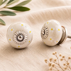 Op de afbeelding: Twee crèmekleurige keramische kastknoppen met een madeliefjespatroon en zilverkleurige metalen accenten. De knoppen hebben een ronde vorm en zijn versierd met kleine gele en witte bloemen. Eén knop is bevestigd aan een schroef.