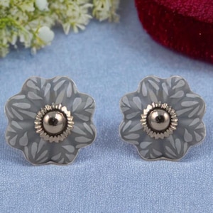 Può includere: Un paio di orecchini d'argento a forma di fiore con un motivo bianco e grigio e un centro dorato.