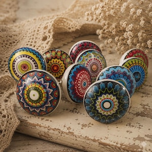 May include: A collection of colorful ceramic cabinet knobs with intricate mandala designs. Each knob features a unique pattern in shades of blue, red, yellow, and green, with white bases. The knobs are arranged on a distressed, white wooden surface.