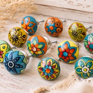May include: A collection of ten round ceramic cabinet knobs featuring hand-painted floral patterns in vibrant colours including orange, yellow, blue, green, and red. Each knob has a silver-toned metal post extending from the back for mounting.