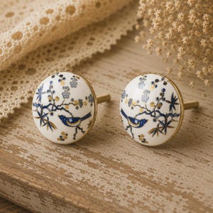May include: A pair of ceramic cabinet knobs. Each knob has a white base, decorated with a blue bird on a branch with blue and gold floral details. They have brass stems and are set on a wooden surface.