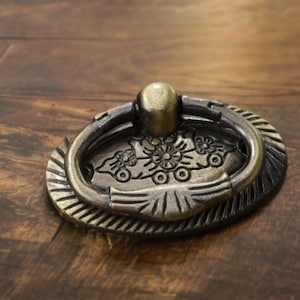 Può includere: Pomello per cassetto in ottone antico con un motivo floreale decorativo. Il pomello presenta una maniglia ad anello e una piastra posteriore ovale con dettagli intricati. L'hardware è posto su una superficie di legno scuro.