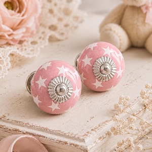 Puede incluir: Dos pomos de armario rosas con estrellas blancas y herrajes plateados. Los pomos son redondos y se asientan sobre una superficie blanca desgastada. Un osito de peluche y una rosa rosa están en el fondo, sugiriendo un tema de habitación infantil.
