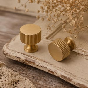 Op de afbeelding: Twee cilindervormige kastknoppen van geborsteld messing. Elke knop heeft een plat, rond bovenoppervlak en een verticaal geribbelde of gegroefde zijtextuur, verbonden met een kleine ronde voet voor bevestiging aan een kast of lade.