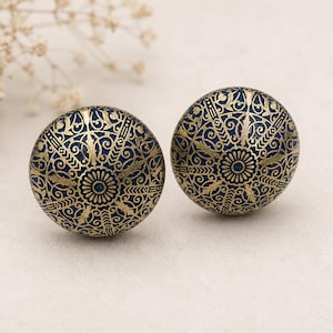 May include: Two round cabinet knobs with a dark blue base and an intricate gold filigree pattern. The design includes a central floral motif and swirling patterns. The knobs are likely made of metal and are suitable for furniture.