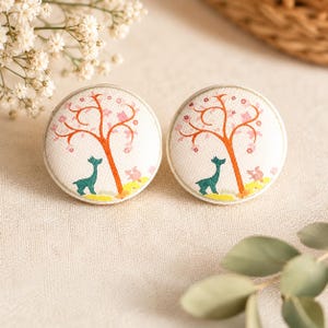 Puede incluir: Dos botones redondos cubiertos de tela blanca. Cada botón presenta un árbol naranja estilizado con flores rosas, un ciervo verde azulado y detalles de suelo amarillos y rosas. Los botones están sobre un fondo neutro, lo que sugiere un artículo de artesanía o un accesorio.