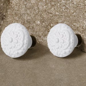Puede incluir: Dos pomos de armario de cerámica blanca con un diseño floral detallado. Los pomos tienen forma redondeada y están montados sobre herrajes metálicos oscuros. Los pomos están sobre un fondo neutro.