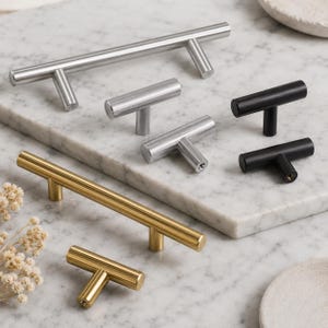 May include: A selection of cabinet handles in silver, gold, and black finishes. The T-bar and bar pulls are displayed on a marble surface, highlighting their various sizes and styles. These handles are suitable for furniture and cabinetry.