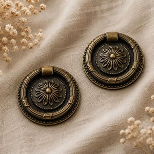 May include: Two antique brass drawer pulls with a floral design. Each pull features a circular shape with a central flower and a ring of decorative details. The pulls are set against a neutral fabric background, suggesting a vintage or rustic aesthetic.