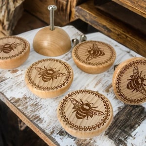 Peut inclure: Ensemble de boutons de meubles en bois et une cheville en bois. Chaque bouton présente un motif d'abeille gravé au laser, certains avec une bordure de feuilles décorative. Les boutons sont brun clair et semblent être en bois naturel.