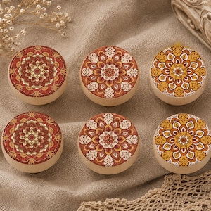 May include: Six circular wooden coasters featuring mandala patterns in red, orange, and cream. They are displayed on a beige linen cloth, with decorative items in the background.
