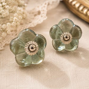 Puede incluir: Dos pomos de armario con forma de flor, hechos de vidrio transparente de color verde pálido con centros de metal plateado. Están sobre una tela color crema, con un ribete de encaje y un espejo con marco dorado en el fondo.