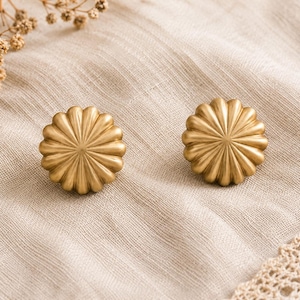 Puede incluir: Dos pendientes de botón dorados con forma de flor. Los pendientes tienen un diseño detallado y acanalado y están colocados sobre una tela de lino beige. También se ven acentos florales secos y un ribete de encaje.
