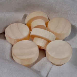 Op de afbeelding: Een verzameling onafgewerkte houten flessenstoppers. De stoppers zijn cilindrisch met een platte bovenkant en een taps toelopende basis. De houtnerf is zichtbaar en de kleur is een lichte, natuurlijke tint. Ze zijn gerangschikt op een grijze stof.