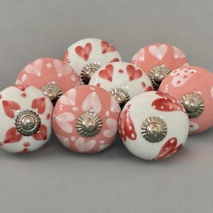 Puede incluir: Un conjunto de ocho pomos de cerámica para armarios. Los pomos redondos tienen un acabado brillante y presentan patrones de corazones rojos sobre fondos blancos y rosas. Cada pomo tiene una base metálica plateada.