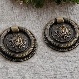 May include: Two antique bronze cabinet pulls with a floral design. Each pull features a circular shape with a central flower, surrounded by a beaded border and a handle. The pulls are made of metal and have a vintage aesthetic.