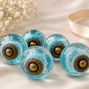 Op de afbeelding: Vijf helderblauwe glazen kastknoppen met een bronskleurig midden en een decoratief ontwerp. De knoppen zijn gerangschikt op een wit oppervlak met een lint en witte bloemen op de achtergrond.