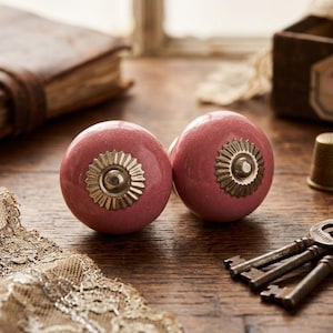 Può includere: Due pomelli per armadi in ceramica rosa con dettagli in metallo color oro. I pomelli sono rotondi con un design floreale. Un set di chiavi antiche, un ditale e un libro vintage sono visibili su una superficie di legno.