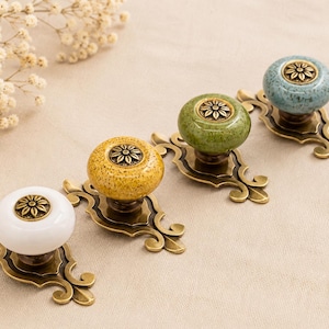 Puede incluir: Cuatro tiradores decorativos para armarios con tapas de cerámica en blanco, amarillo, verde y azul. Cada tirador presenta un diseño floral y está montado sobre una base ornamental de color latón antiguo. Los tiradores están dispuestos sobre una superficie de tela de tono neutro.