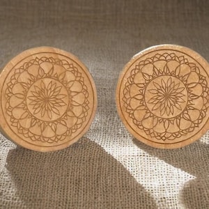 Peut inclure: Deux boutons de meuble ronds en bois brun clair. Chaque bouton présente un motif de mandala floral gravé et détaillé. Les boutons sont présentés sur une surface texturée de couleur neutre, avec des ombres ajoutant de la profondeur.