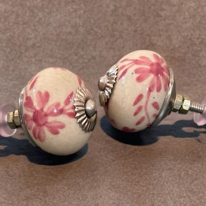 May include: Two decorative cabinet knobs. Each knob features a cream-colored ceramic sphere with a pink floral design. The knobs have silver-colored metal accents and are attached to a screw with a nut and washer.