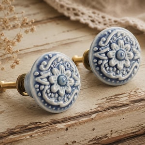 May include: Two blue and white ceramic cabinet knobs with a floral design. The knobs have a brass-coloured base and are set against a rustic, light-coloured wooden surface. Decorative furniture hardware.