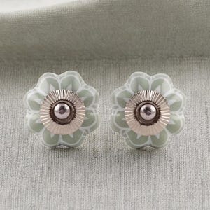 Op de afbeelding: Twee bloemvormige kastknoppen. De knoppen hebben een lichtgroene basis met een wit bloemmotief. Het midden is zilverkleurig met een kleine, ronde, zilveren knop. De knoppen staan op een lichtgroene achtergrond.