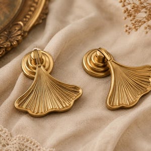 May include: Two gold cabinet pulls featuring a ginkgo leaf design. The metal pulls have a circular base and detailed veining on the leaf design. They are displayed on a beige fabric. The pulls are likely for furniture.