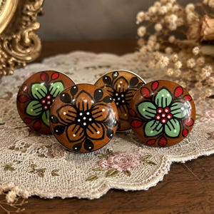 Op de afbeelding: Vier ronde houten kastknoppen met een bruine basis en bloemmotieven in groen, rood en zwart. De knoppen liggen op een kanten kleedje, wat een vintage uitstraling geeft. Decoratieve meubelbeslag.