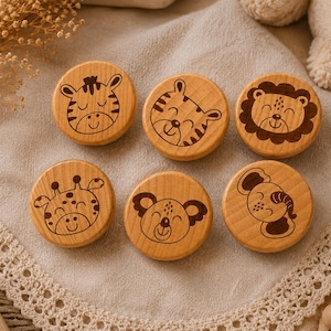 May include: Six wooden discs, each with a different smiling animal face etched in brown. Animals include a zebra, tiger, lion, giraffe, koala, and elephant. The discs are arranged on a beige cloth. The wood is light-coloured.