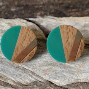 May include: Two round stud earrings. Each earring features a teal green circle paired with a section of wood grain and a copper-colored accent. The earrings are set against a neutral background.
