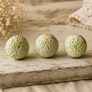May include: Three round, light green ceramic knobs with a raised leaf pattern. The knobs are arranged in a row on a distressed, white-painted wooden surface. The background includes dried flowers and a lace cloth.