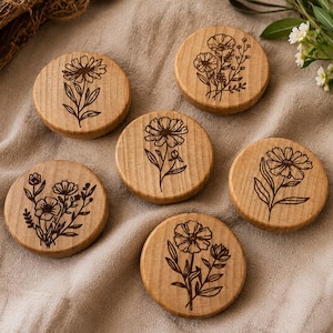 Puede incluir: Seis posavasos redondos de madera con diseños florales grabados. Los posavasos son de color marrón claro y presentan varias ilustraciones botánicas. Están dispuestos sobre una superficie de lino beige, con vegetación en el fondo.