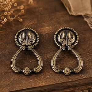 May include: A pair of antique brass drawer pulls with intricate floral and leaf patterns. Each pull has a circular top with detailed carvings and a teardrop-shaped handle. They are displayed on a dark wooden surface.