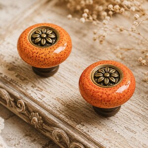 Könnte beinhalten: Zwei orangefarbene Schrankknöpfe mit gesprenkelter Oberfläche und einem bronzefarbenen Blumendesign. Die Knöpfe sitzen auf einem dunkelbraunen Sockel. Die Knöpfe werden auf einer verwitterten Holzoberfläche präsentiert.