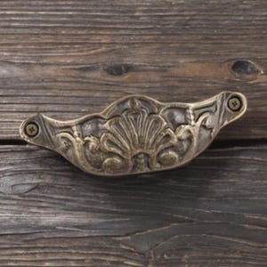 May include: An antique brass drawer pull with an ornate shell design, fixed to a weathered wooden surface. The pull is secured with two screws and features detailed floral and scrollwork patterns. A decorative hardware element.