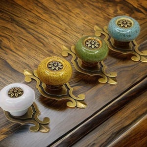 Pode incluir: Quatro puxadores decorativos para armários em branco, amarelo, verde e azul. Cada puxador tem um design floral e é montado numa placa traseira ornamental de cor latão. Os puxadores estão dispostos numa superfície de madeira.