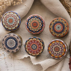 Peut inclure: Six boîtes rondes décoratives avec des motifs de mandala complexes dans diverses couleurs, dont le bleu, l'orange et le rose. Les boîtes sont disposées sur un tissu de couleur neutre, mettant en valeur leurs motifs détaillés.