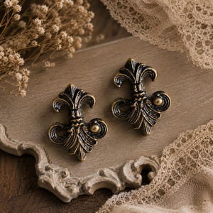 May include: Two antique bronze-coloured fleur-de-lis cabinet knobs. The knobs feature detailed designs and a vintage appearance. They are presented on a wooden surface with decorative elements, indicating a focus on home decor and hardware.