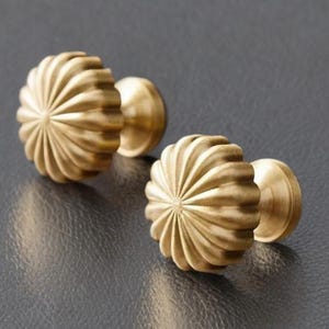 May include: Two brass cabinet knobs with a fluted, sunburst design. The knobs have a warm, golden hue and a smooth, polished finish. The hardware is designed for use on drawers or cabinets.