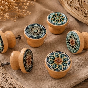 May include: Six wooden cabinet knobs with decorative floral designs. The knobs have a light wood base and a round, patterned top with teal, orange, and gold accents. The knobs are arranged on a tan fabric surface, suitable for drawers or cupboards.