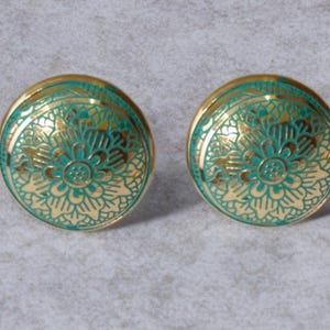 Puede incluir: Dos pomos de armario redondos dorados con un diseño floral turquesa. Los pomos tienen un acabado metálico y un patrón en relieve detallado. El color turquesa se aplica a las zonas hundidas del diseño, creando contraste.