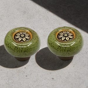 May include: Two green ceramic cabinet knobs with a speckled finish. Each knob has a gold-coloured rim and a floral design in the centre. These knobs are likely for furniture or decorative use.