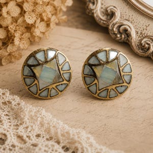 May include: Two circular cabinet knobs with a brass finish. Each knob has a central square and surrounding triangles of iridescent, pale blue and cream mosaic tiles. The knobs are displayed on a neutral, vintage-style background.