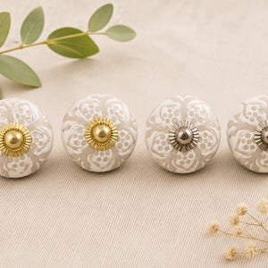 May include: Four decorative ceramic cabinet knobs. Each knob is white with a grey floral pattern and a metallic centre. Two knobs have gold centres, and two have silver centres. The knobs are arranged in a row on a neutral surface.