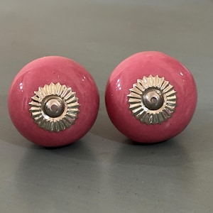 Könnte beinhalten: Zwei runde, rosa Keramik-Schrankknöpfe mit silberfarbenen Metallakzenten. Die Knöpfe haben eine glänzende Oberfläche und ein dekoratives, sonnenstrahlenartiges Design um das zentrale Schraubenloch. Geeignet für Möbel oder Heimdekoration.